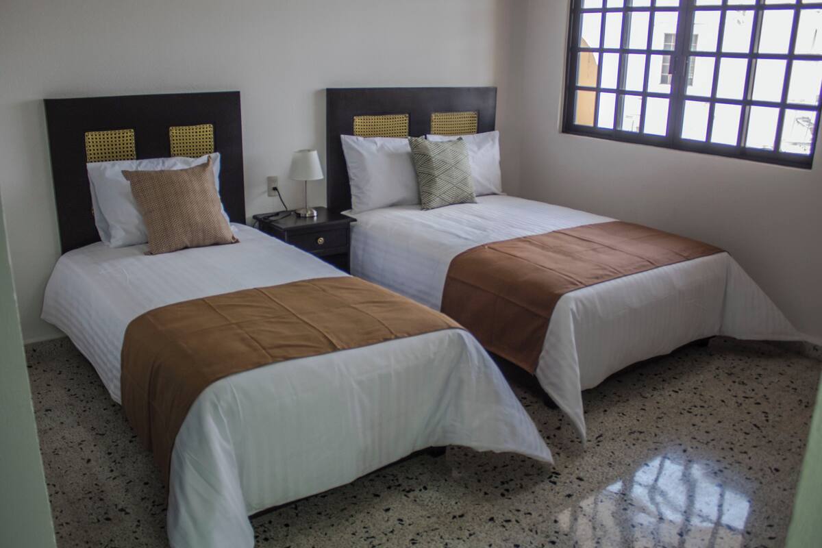 A hotel room with two single beds, each with a headboard, a nightstand, a lamp, and a window with a view.