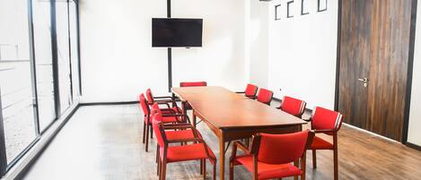 Un salón de conferencias con una larga mesa y sillas rojas, un televisor de pantalla plana en la pared y ventanas amplias.