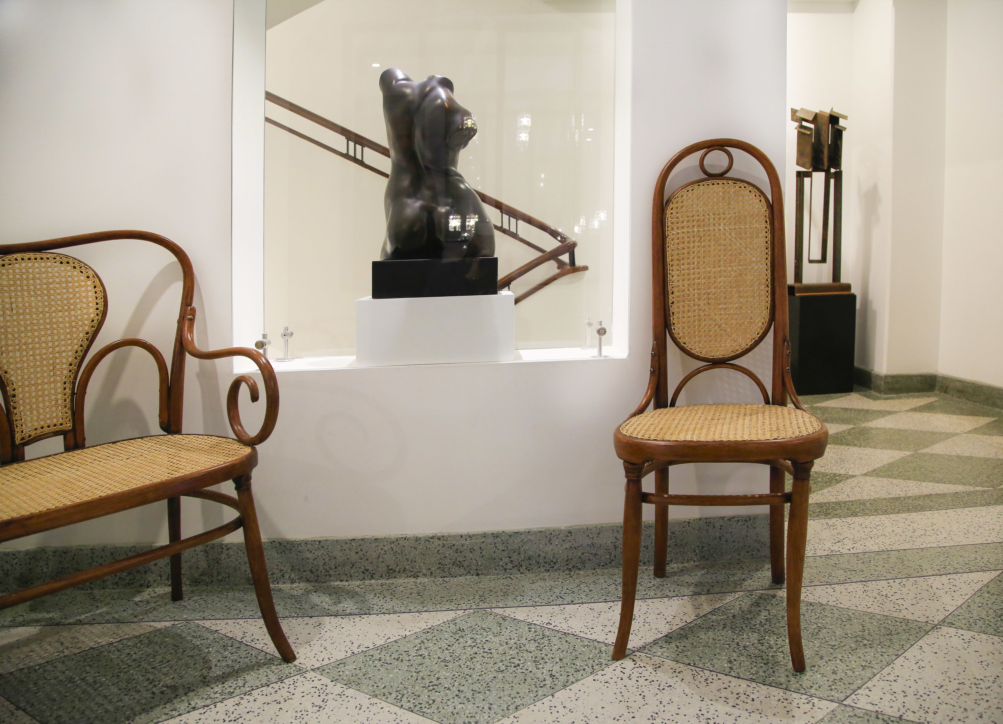 Une pièce comprenant deux chaises en bois à assise tressée, un mur blanc et une fenêtre donnant sur une sculpture.