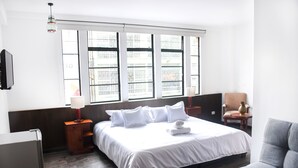 A hotel room with a large bed, wooden flooring, a window with blinds, a bedside table with a lamp, and a chair. - Hotel Museo San Moritz (Bogotá)