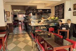 A café with a checkered floor, red chairs, a glass display case with food items, and a chalkboard menu. - Hotel Museo San Moritz (Bogotá)