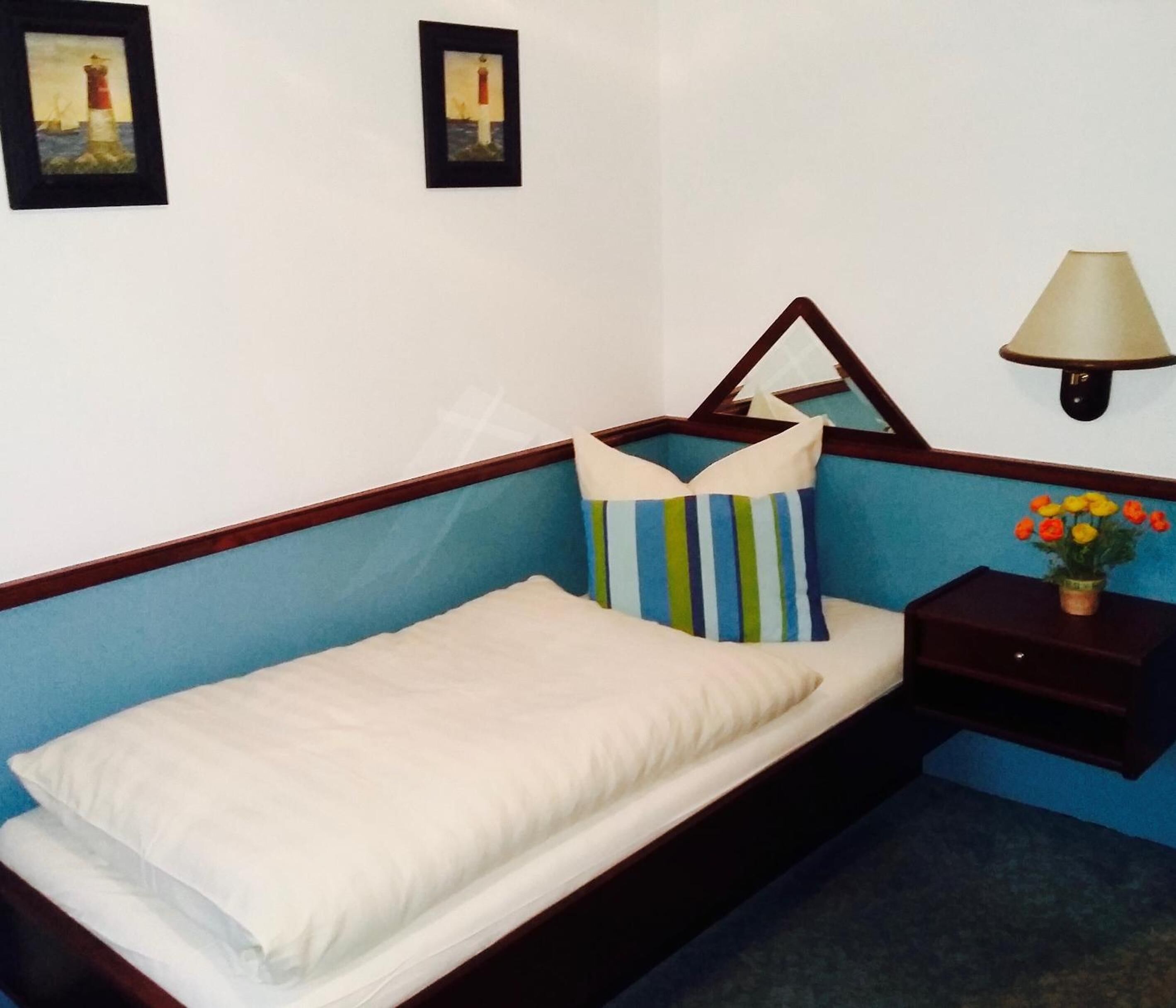 A single bed with a wooden frame, a pillow, a bedside table with a lamp, and a small vase with flowers.