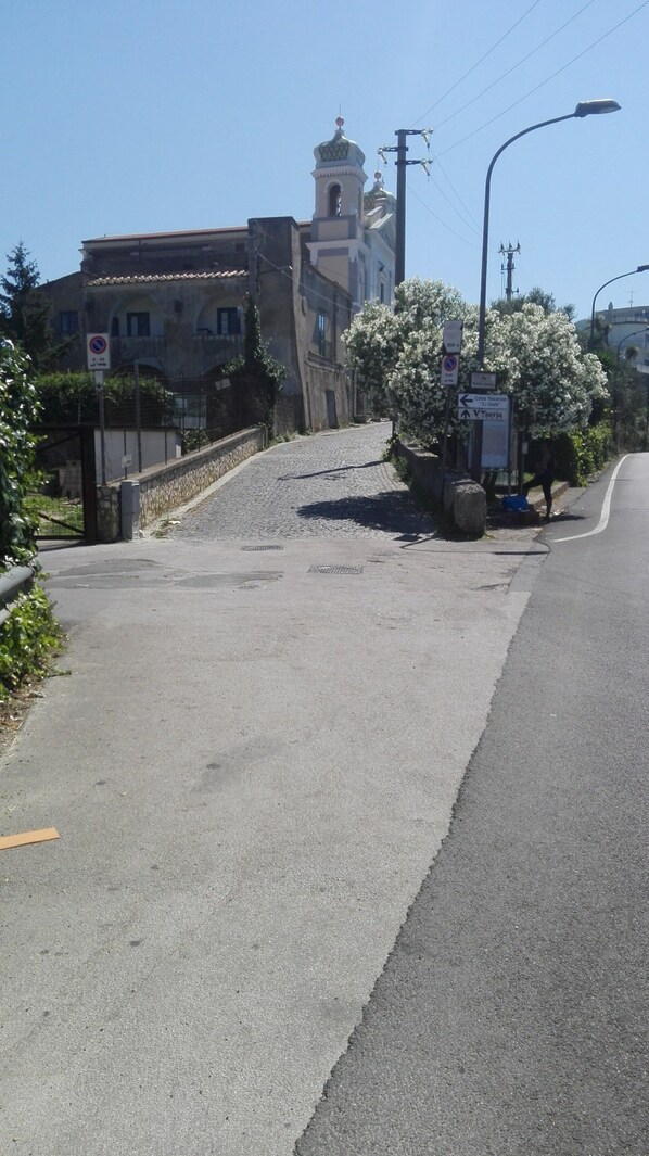 Una carretera pavimentada con una escalera de piedra que conduce a un edificio con una torre de campanas. Hay señales de calle y un poste de electricidad con cables.