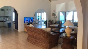 A spacious living area with a television, wicker furniture, a lamp, and a view of the kitchen and balcony. - Patricks Bay Villa (Island Harbour)