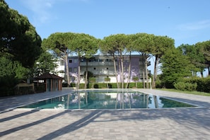 Una piscina con bordo piastrellato, circondata da alberi e un edificio con balconi.
