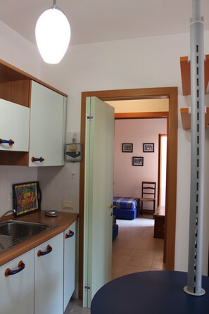 Una cucina con mobili bianchi, un lavello e un piano di lavoro blu. Si apre una porta su un soggiorno con un divano blu e una sedia in legno.