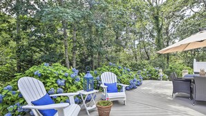 A wooden deck with white Adirondack chairs and a table, surrounded by lush greenery and blue hydrangeas. - ~ 4 Mi to Dtwn: Chatham Home w/ Fire Pit (South Chatham)