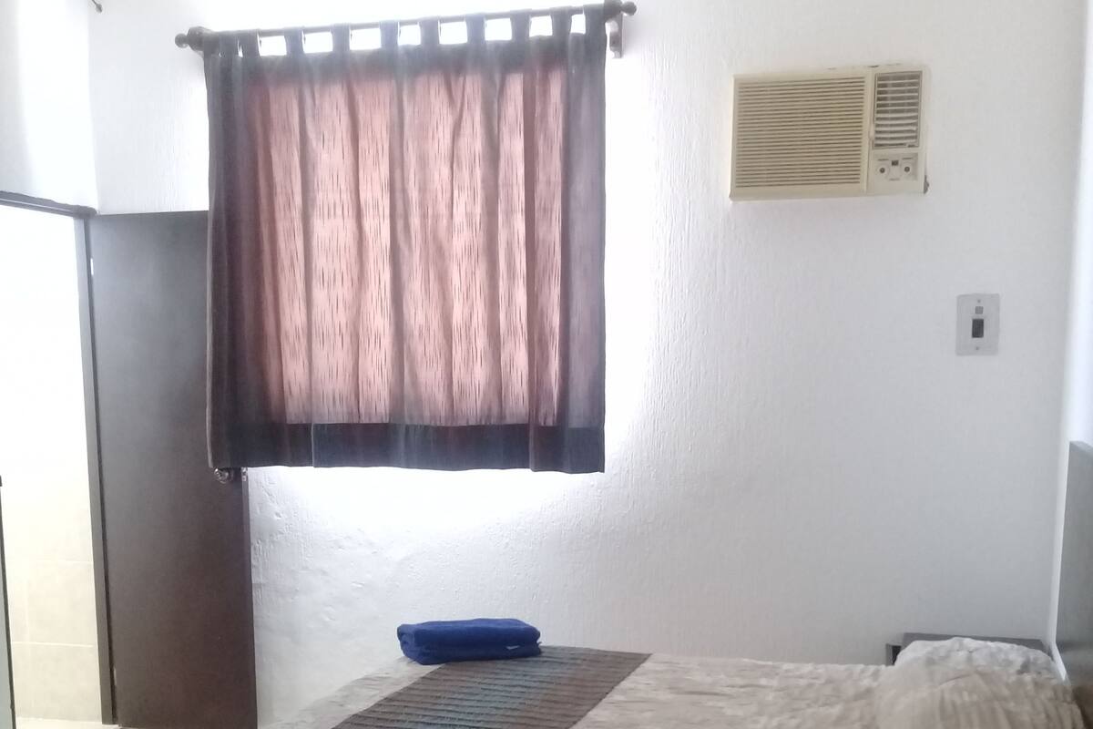 Una habitación con cama, cortinas en la ventana, aire acondicionado de pared y un mando a distancia sobre la mesa, además de una toalla en la cama.