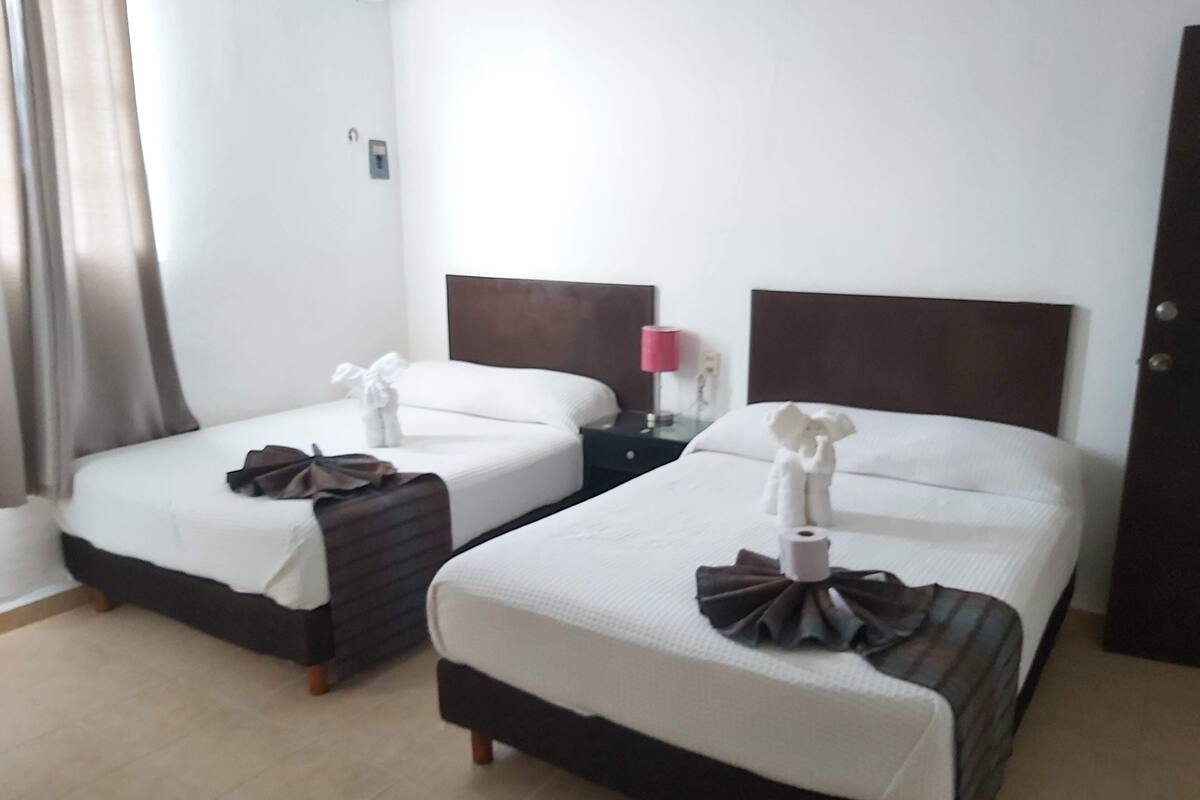 Habitación de hotel con dos camas, cada una con una colcha blanca y un adorno marrón sobre la cama. Entre las camas, un mesita de noche con una lámpara.