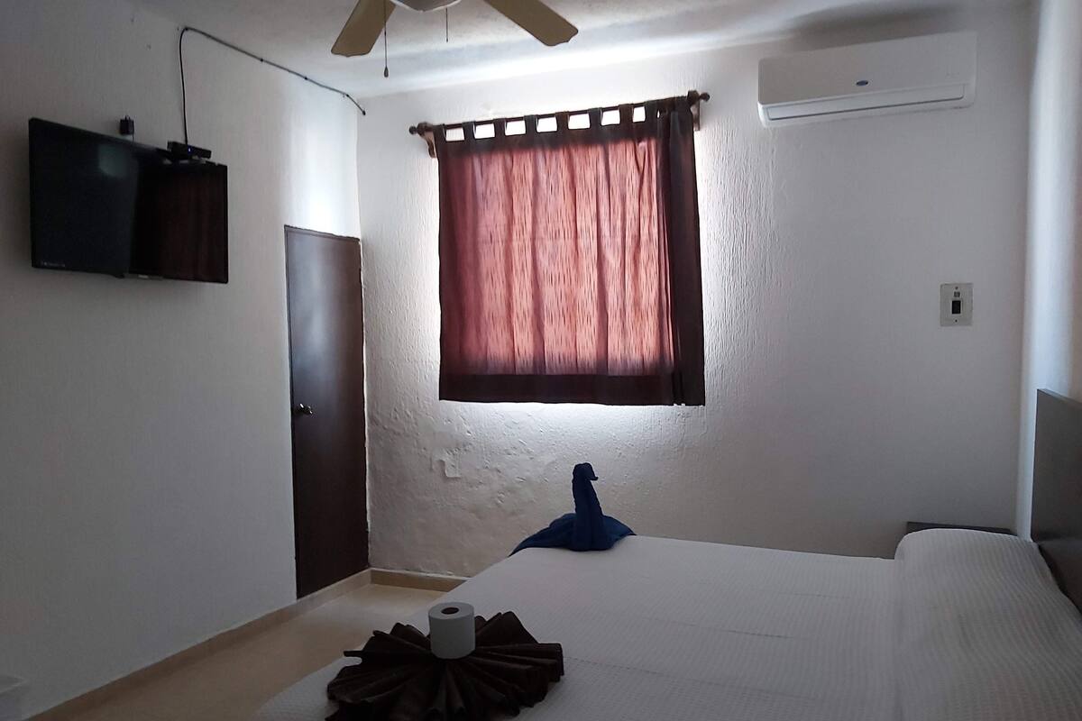 Un dormitorio con cama, ventilador de techo, televisor montado en la pared y ventana con cortinas.