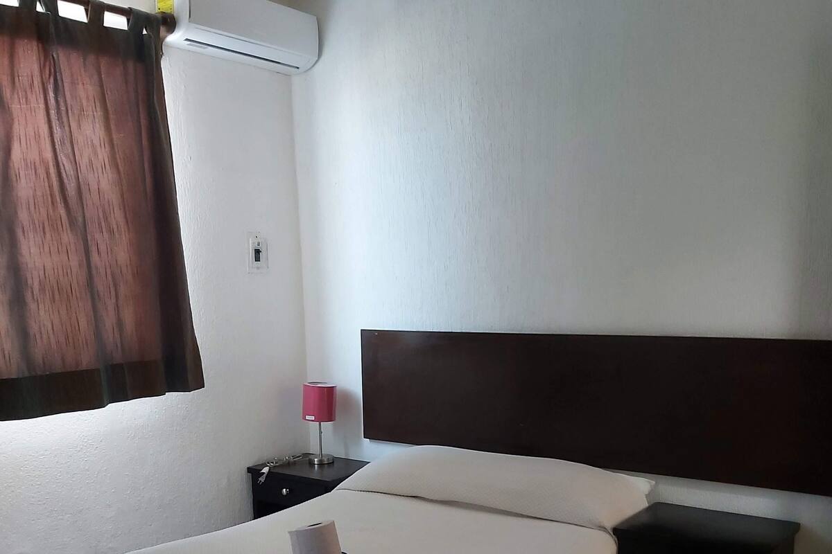 Una cama bien hecha con un colchón blanco y un cabecero oscuro. Junto a la cama, una mesita de noche negra con una lámpara.