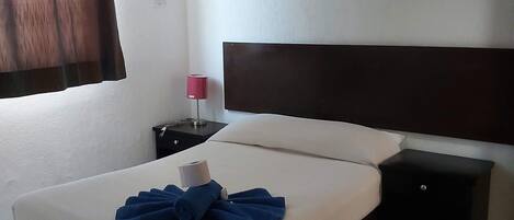 Un lit bien fait, avec un matelas blanc et une tête de lit sombre. Une petite table de chevet noire avec une lampe se trouve à côté du lit.
