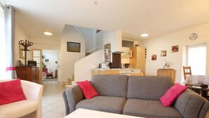 A living room with a grey sofa, a white coffee table, and a red pillow. There is a kitchen in the background with wooden cabinets and a stove.