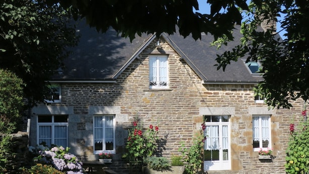 A stone house with a symmetrical facade, two windows with shutters, and a small garden with flowers and shrubs.
