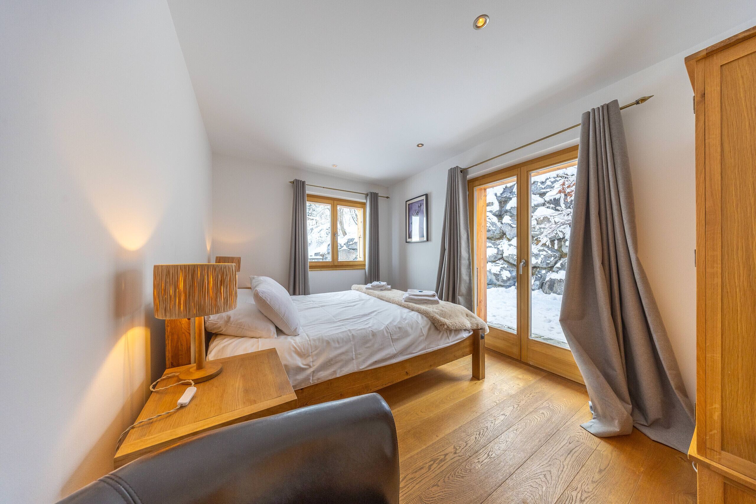 A modern bedroom with a wooden bed, a bedside table with a lamp, a chair, and a large window with curtains.