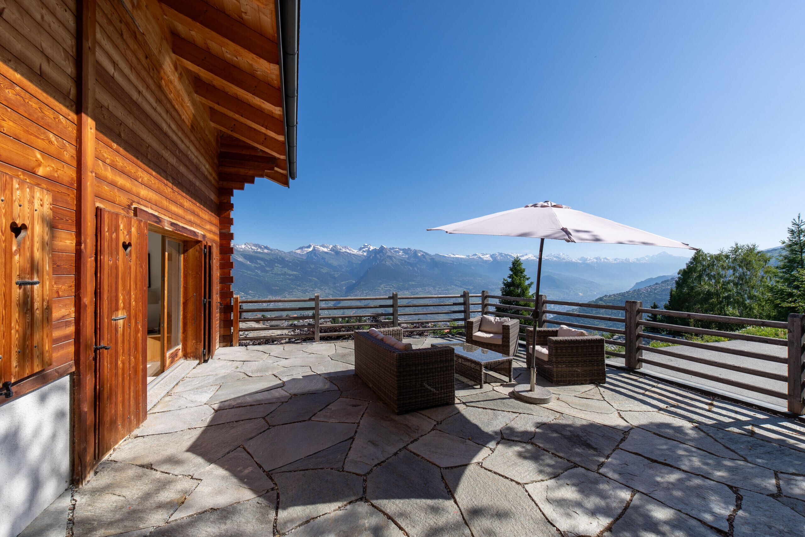 Una cabaña de madera con un patio de piedra, mobiliario exterior y vistas a montañas nevadas.