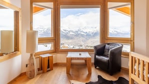 A cozy room with a large window offering a mountain view, a wooden table, a leather chair, and a lamp.