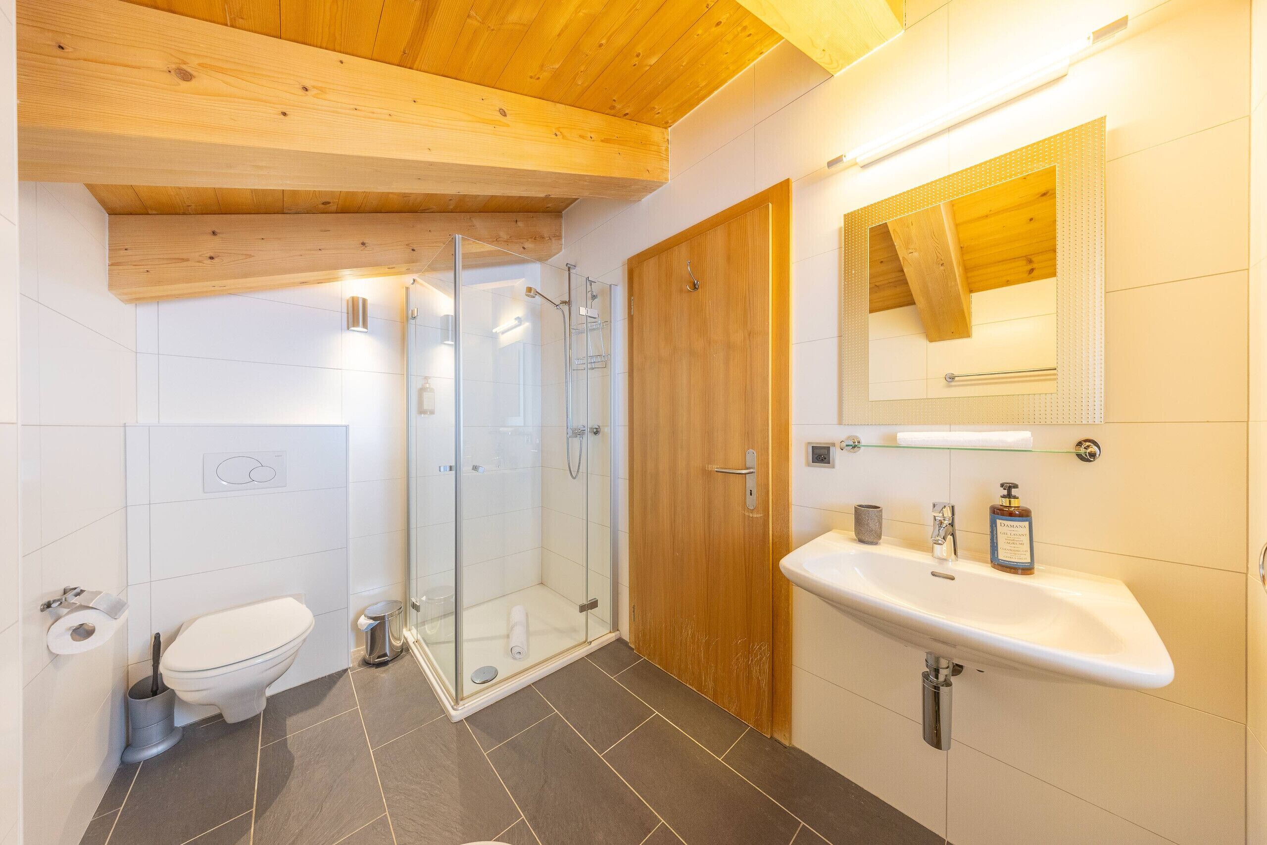 A modern bathroom with a glass shower, wooden ceiling, white tiles, and a wooden door.