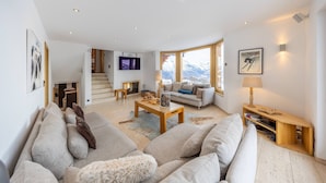 A spacious living area with a sofa set, a coffee table, and a fireplace. There is a television mounted on the wall and a window with a view of mountains.