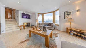 A spacious living area with a wooden coffee table, a sofa, and a fireplace. There is a staircase leading to an upper level, and a large window offering a mountain view.