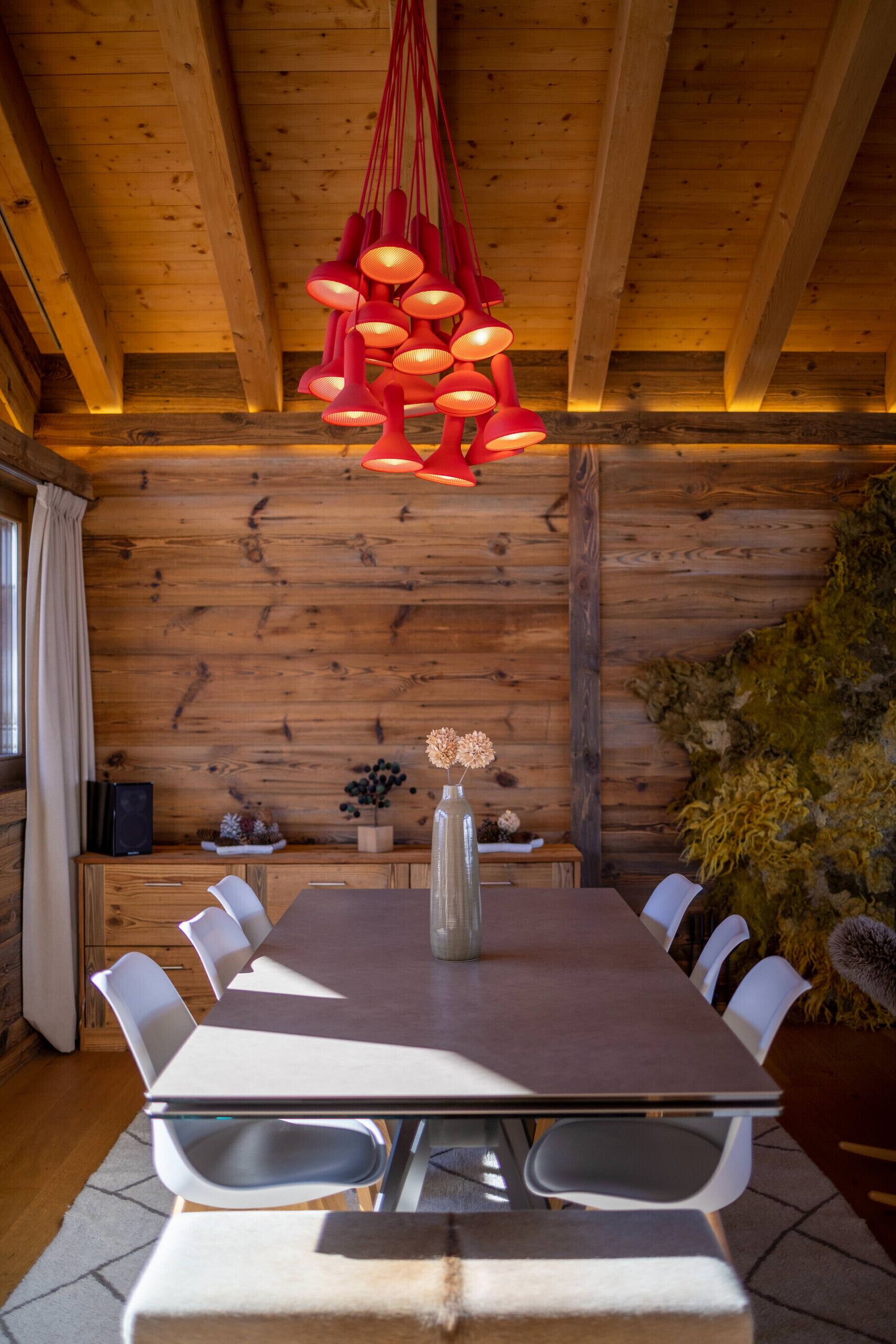A wooden interior with a dining table, chairs, a hanging red light fixture, and a vase with dried flowers.