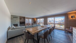 Ein großzügiger Wohnbereich mit einem großen Holztisch, bequemen Sitzgelegenheiten, einem Steinkamin und einem Balkon mit Bergblick.