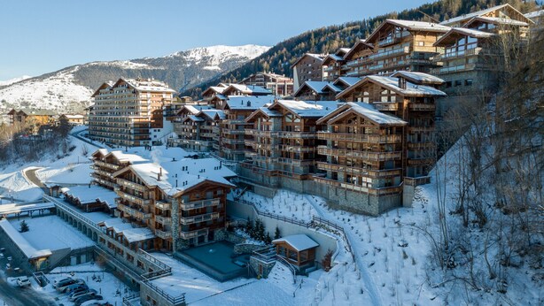 Ein Skigebiet mit mehreren Holzbauten, einem Parkplatz und verschneiter Landschaft.