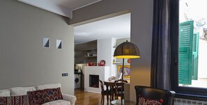 Een moderne woonkamer met een bank, een zwarte lederen fauteuil, een eettafel en een keuken op de achtergrond.
