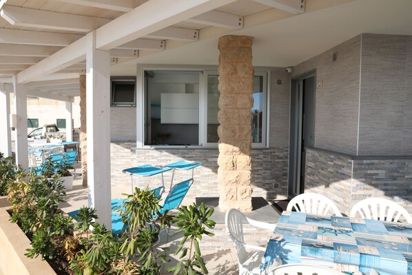 An outdoor patio area with white chairs and blue umbrellas, a stone pillar, and a view of the interior through a glass door.