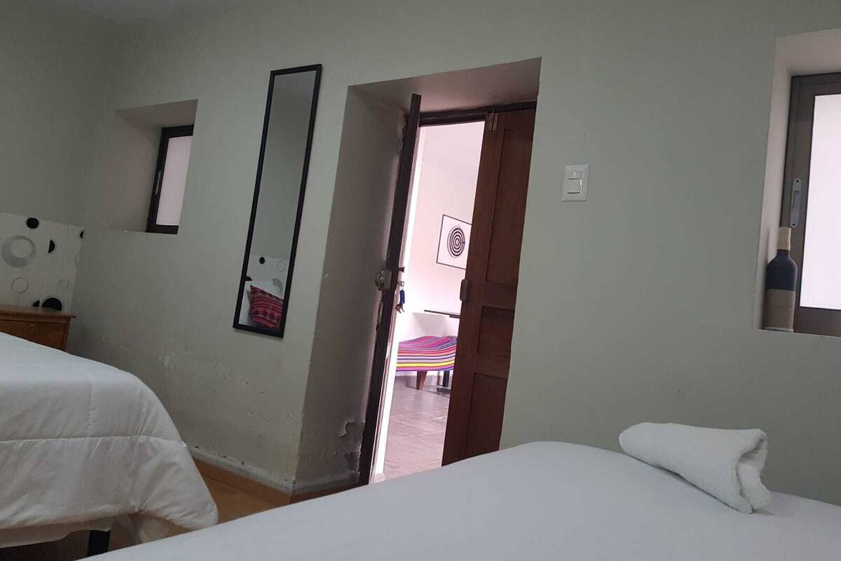 Una habitación de hotel con dos camas, un espejo y una puerta que da a un pasillo con un banco a rayas de colores.