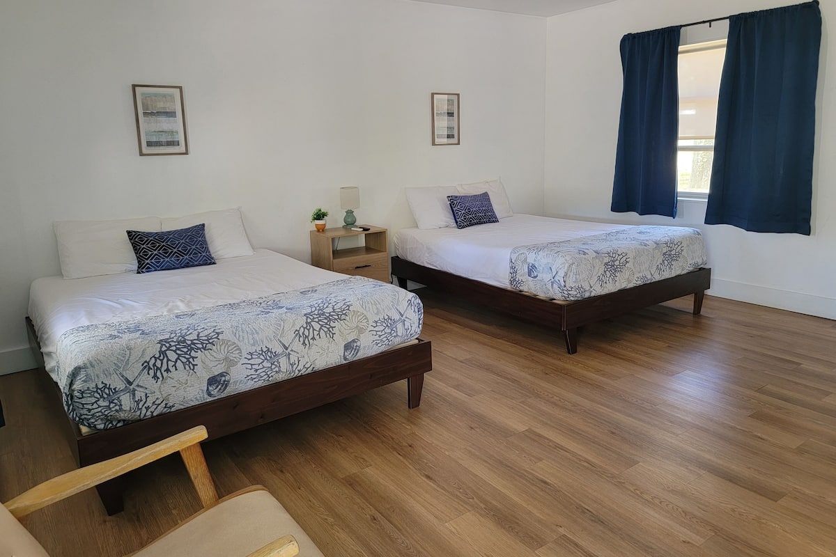 A room with two single beds, wooden flooring, a ceiling fan, and a window with curtains.