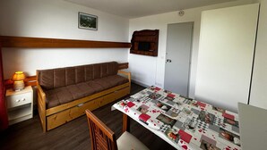 A small, cozy room with a wooden sofa, a small table with a quilt, a wooden chair, a lamp, a picture on the wall, and a door.