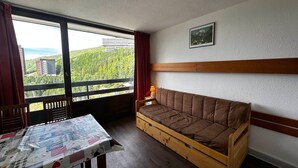 A room with a wooden sofa, a table with a quilt, a window with a view of greenery and buildings, and a framed picture on the wall.