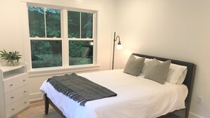 Une chambre à coucher comprenant un lit, une commode, une fenêtre donnant sur un paysage verdoyant et une lampe sur pied.