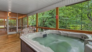 Un bain à remous dans une pièce dotée de grandes fenêtres, d’une terrasse en bois et offrant une vue sur une forêt.