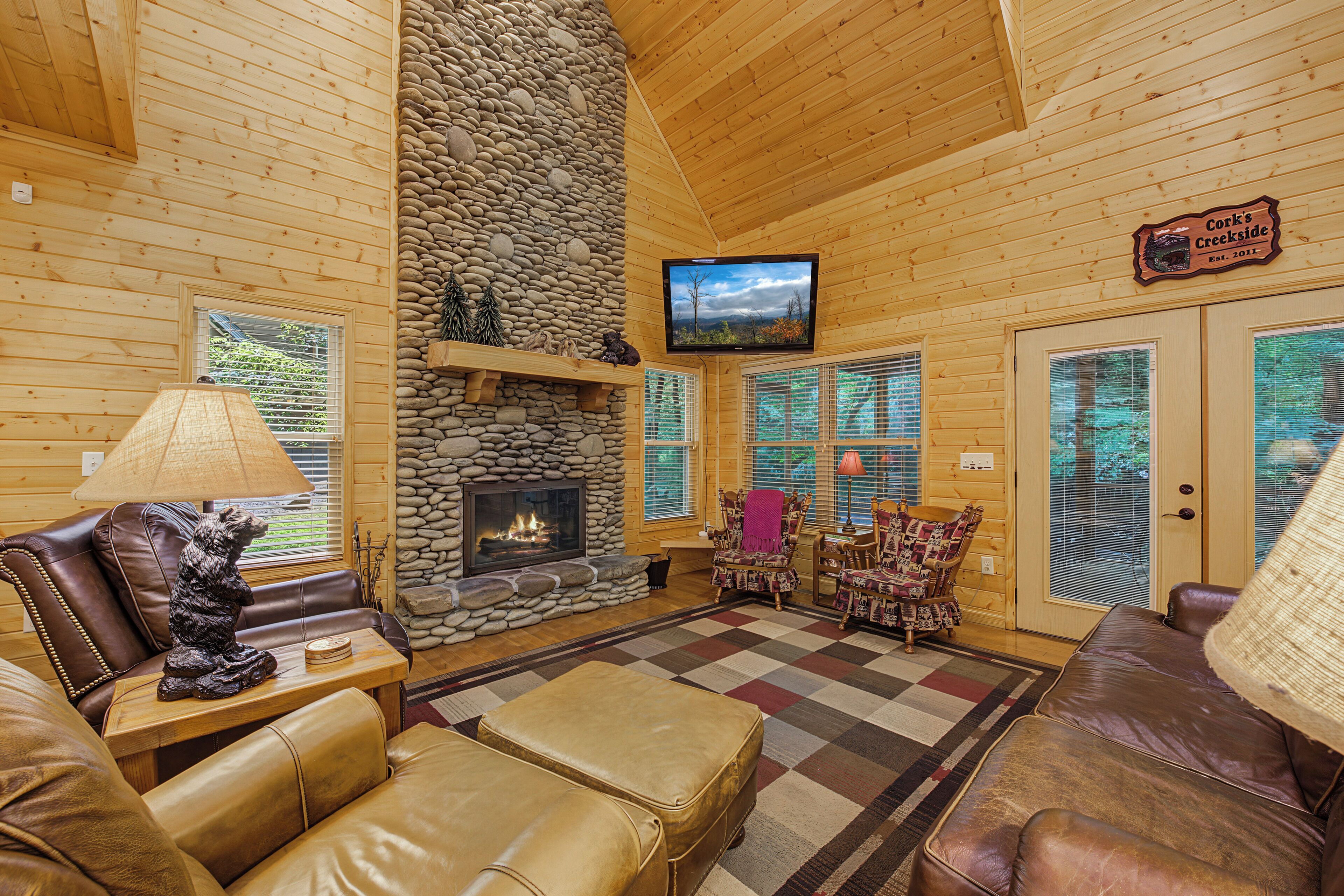 A log cabin interior with a stone fireplace, leather furniture, and a television mounted on the wall.