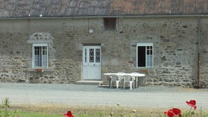 Un bâtiment en pierre avec une porte blanche, deux fenêtres et un petit porche doté d’une table et de chaises.