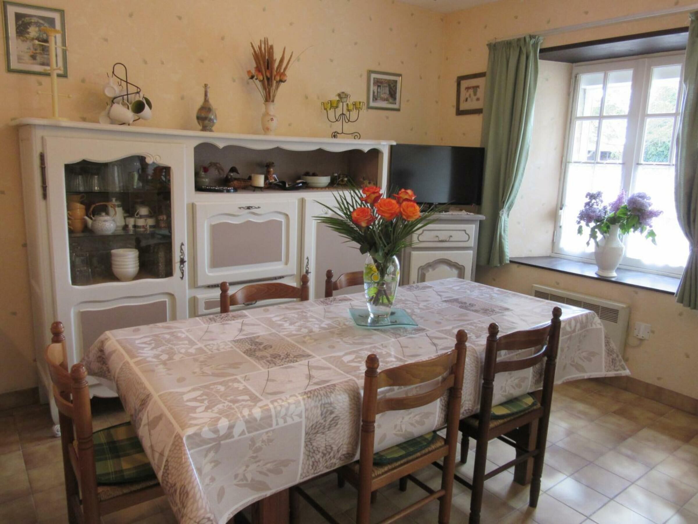 A dining area with a table set for four, surrounded by wooden chairs with green cushions. A vase with orange flowers is on the table.