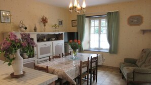 On aperçoit un coin repas avec une table dressée pour quatre personnes, entourée de chaises en bois. Un coin cuisine, avec des placards et un plan de travail, est visible sur la gauche.