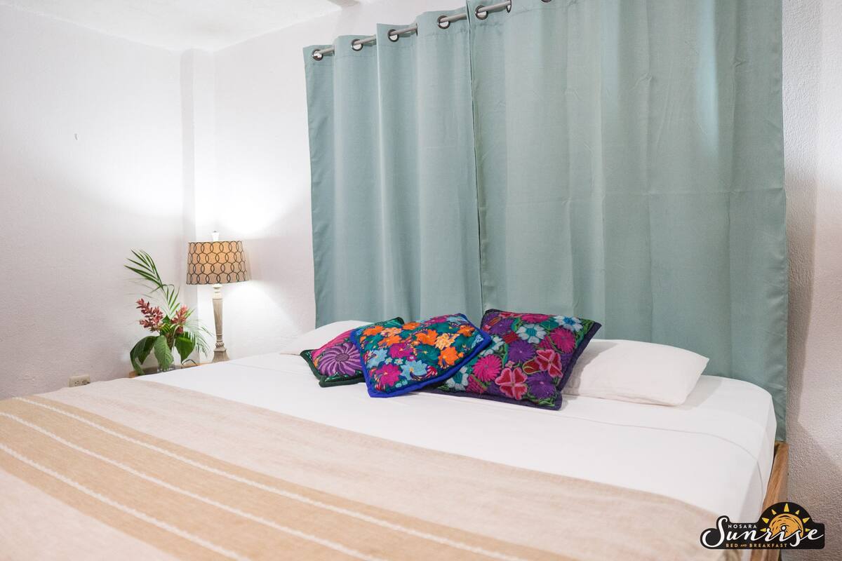 A bed with white bedding, a green curtain, and two colorful pillows.