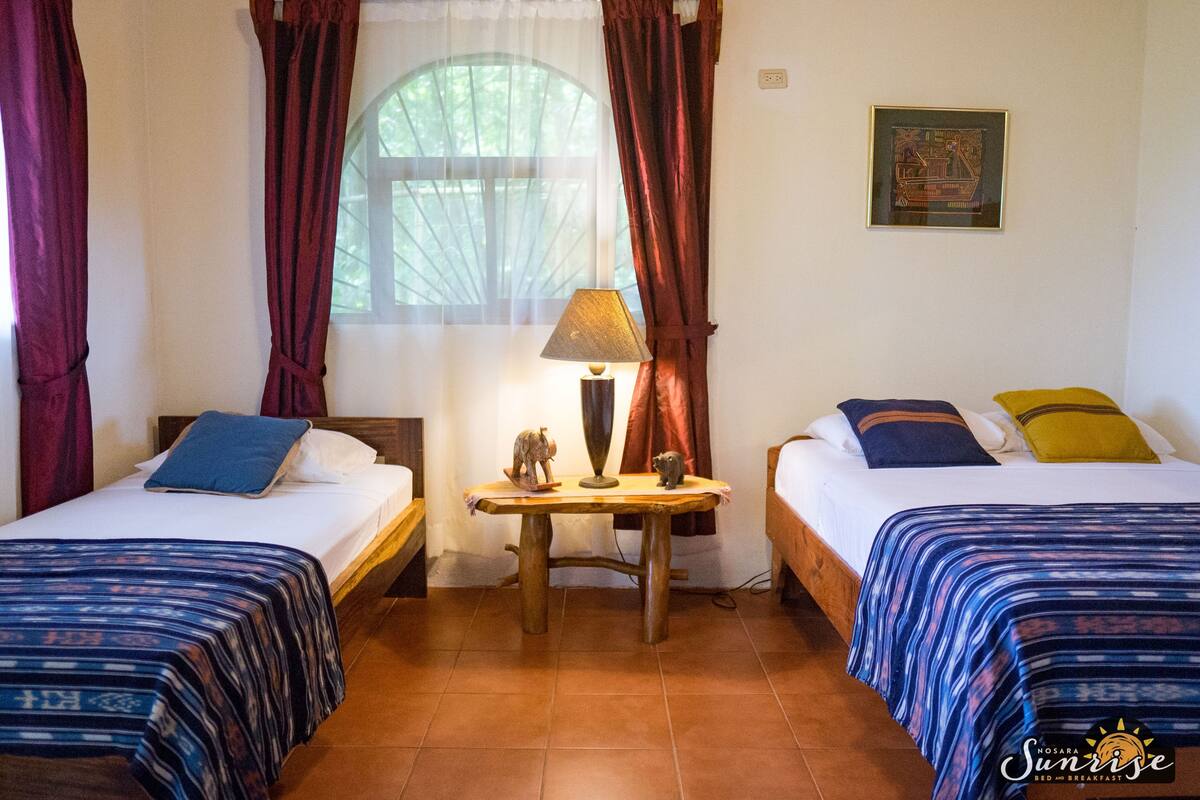 A room with two beds, a small table with a lamp, and a window with curtains.