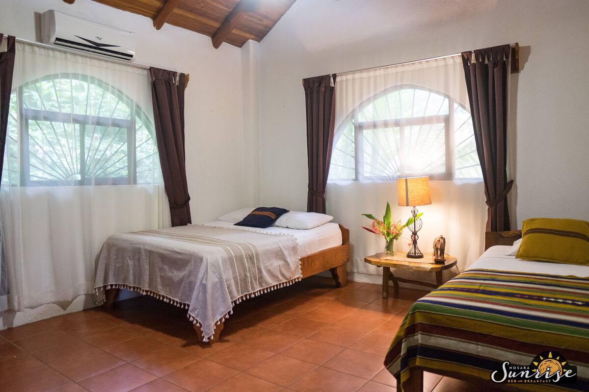 A room with two beds, a small table with a lamp, and a window with curtains.