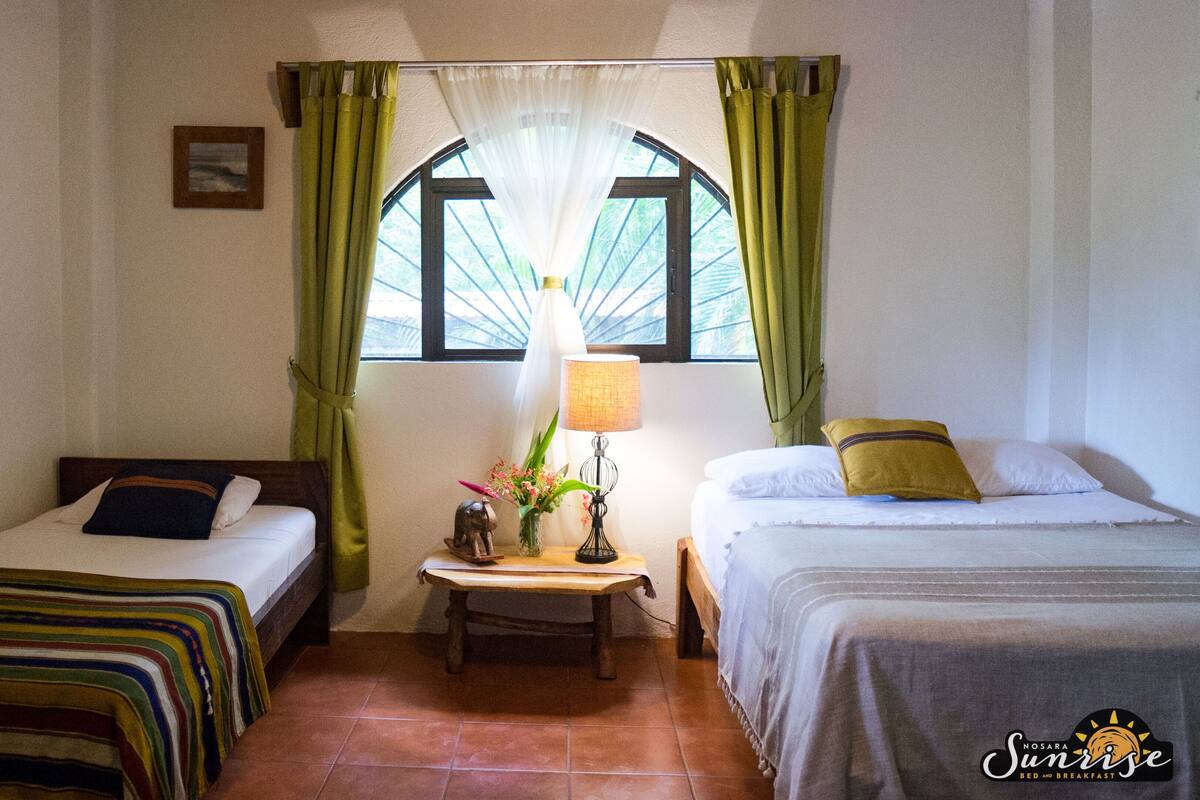 A hotel room with two single beds, a nightstand with a lamp, and a window with green curtains.
