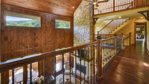A spacious interior with wooden paneling, a stone fireplace, a staircase, and a balcony with a view of the forest. - Fabulous Mountain Views, Secluded, Luxurious , Arcades, Theater, More (Gatlinburg)