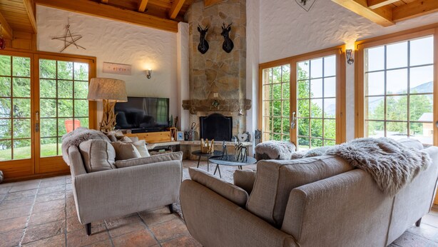 A cozy living room with a fireplace, stone wall, wooden beams, and large windows. - Chalet Pierre Blanche - Ski-in/out (Haute-Nendaz)