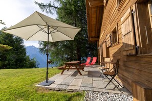 A wooden cabin with a picnic table and chairs, a large umbrella, and a view of mountains. - Chalet Pierre Blanche - Ski-in/out (Haute-Nendaz)