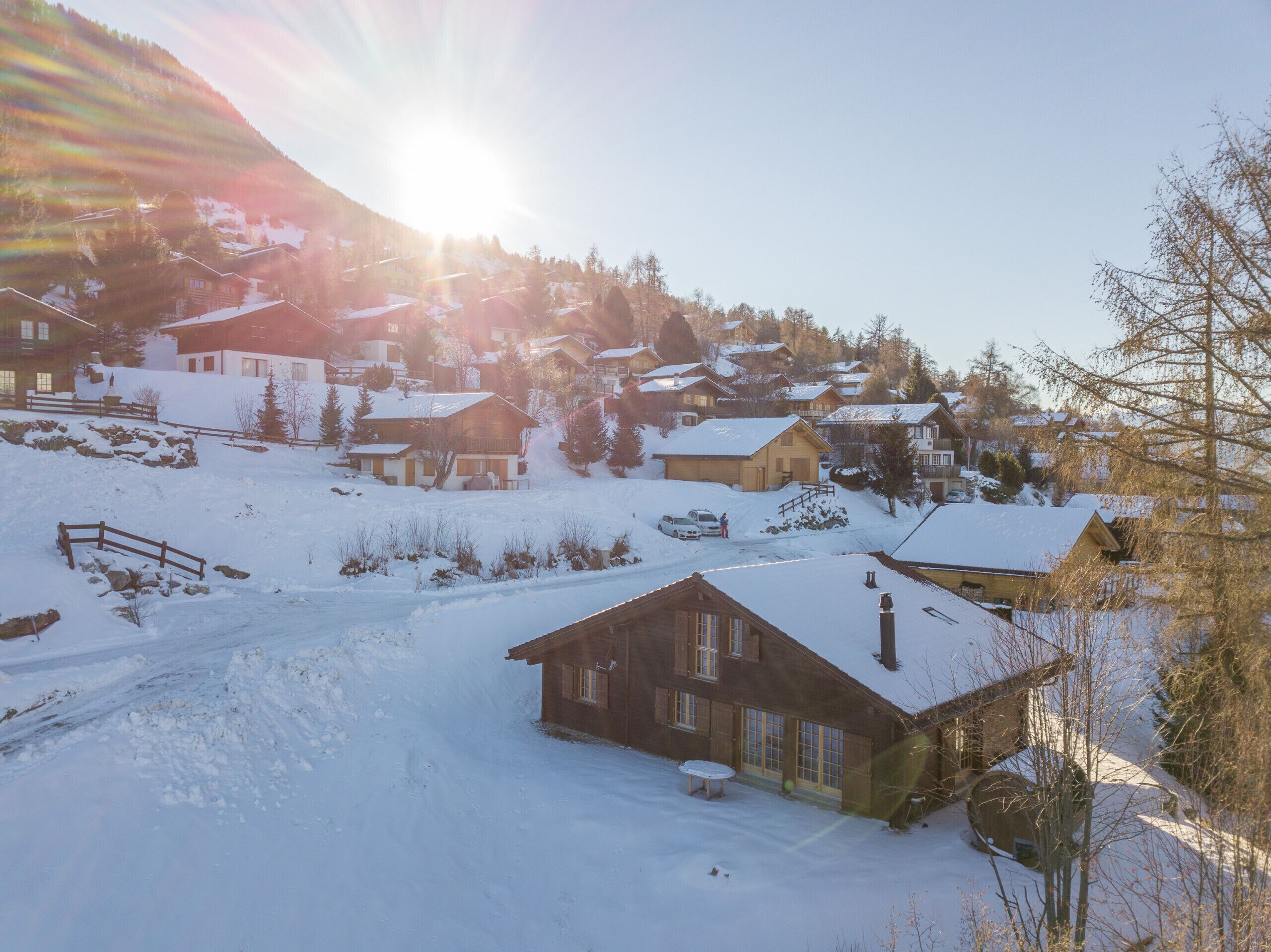 Un village enneigé, avec ses maisons traditionnelles en bois, un ciel dégagé et un soleil brillant derrière les montagnes.