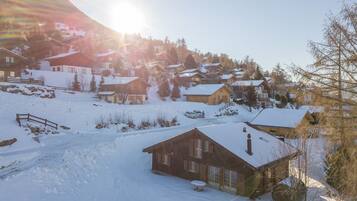 Un village enneigé, avec ses maisons traditionnelles en bois, un ciel dégagé et un soleil brillant derrière les montagnes.