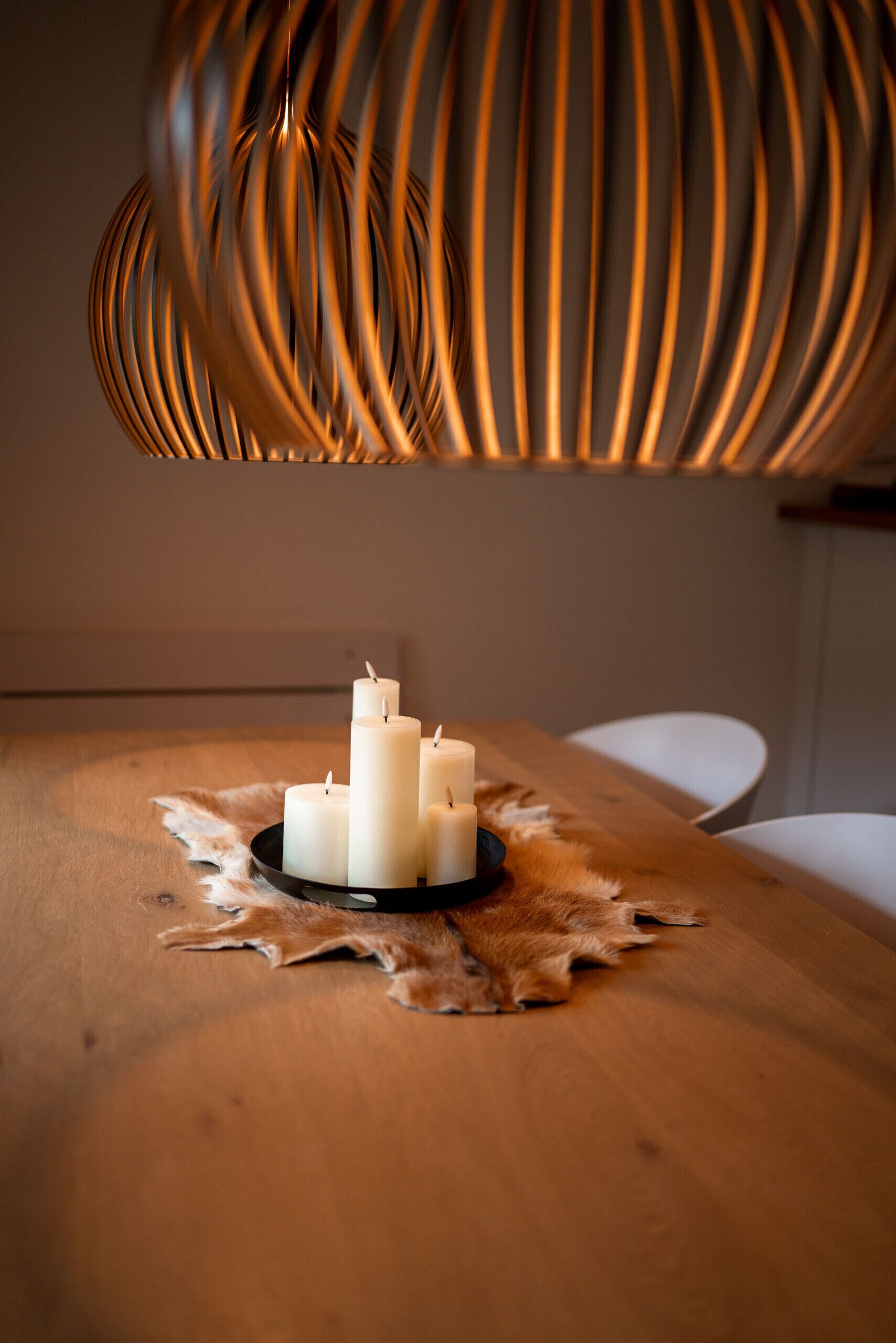 Una mesa de madera con tres velas blancas sobre un posavasos de cuero, bajo una lámpara colgante moderna con textura de nervaduras.