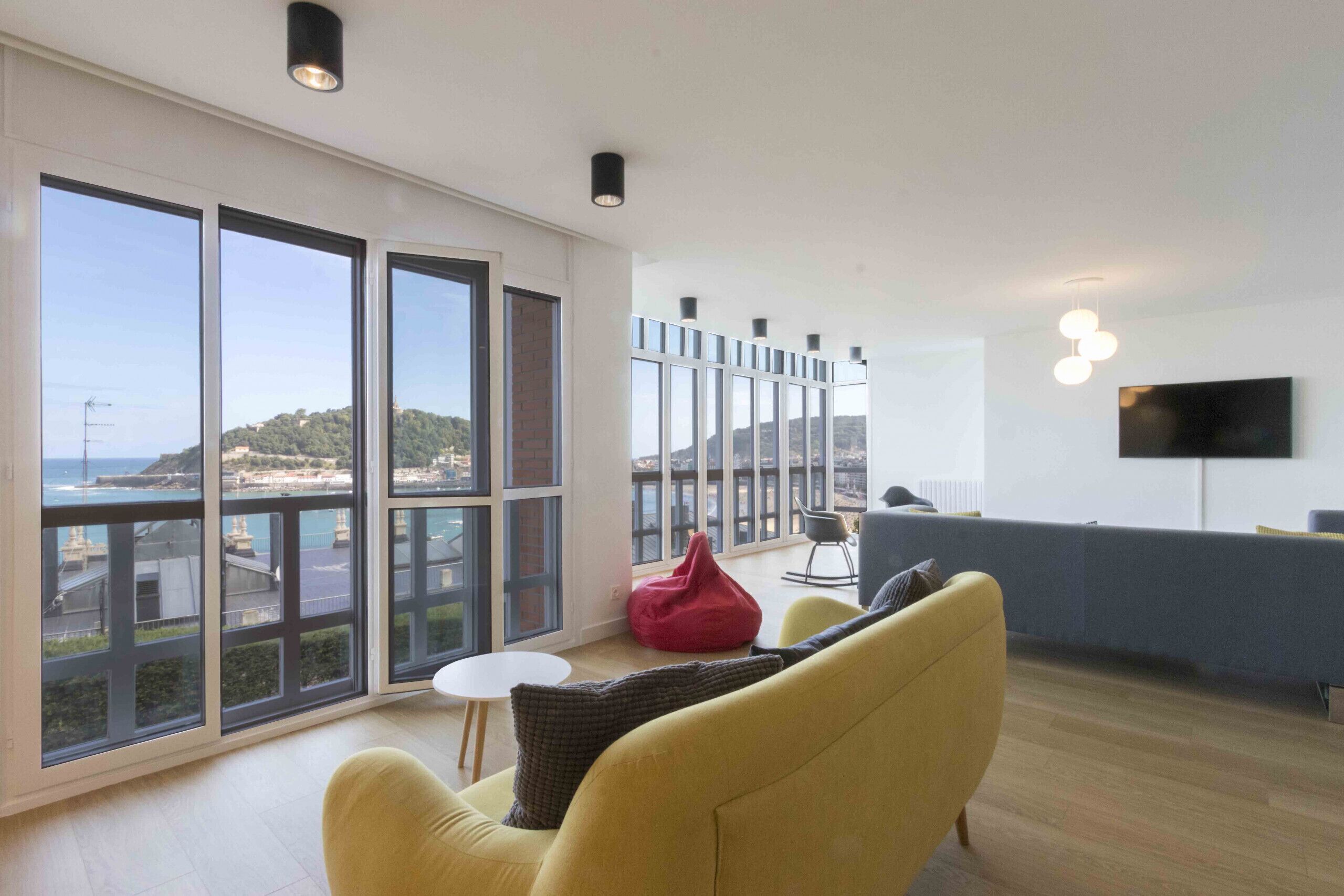 A modern living room with large windows, a yellow sofa, a red bean bag, and a flat-screen TV mounted on the wall.